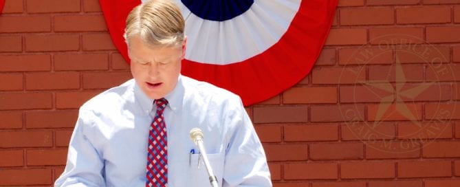 Man in white shirt and red tie speaking at a podium outdoors.