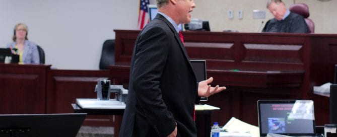 A lawyer in a courtroom, speaking to the judge, with a computer and legal documents on the desk.