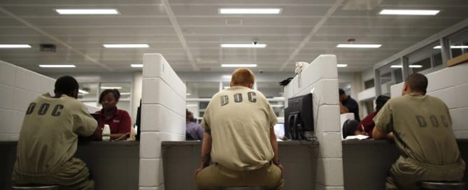 Detainees in orange jumpsuits seated at desks in a detention center.
