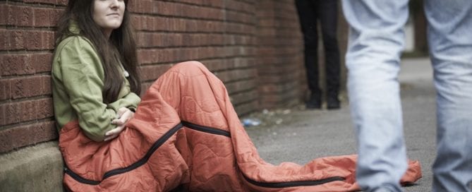 Woman sitting on sidewalk in orange sleeping bag against brick wall, looking distressed.