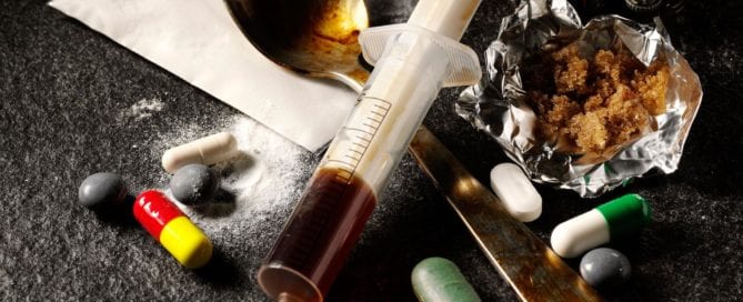 Drug paraphernalia including a syringe, pills, and a spoon on a dark surface.