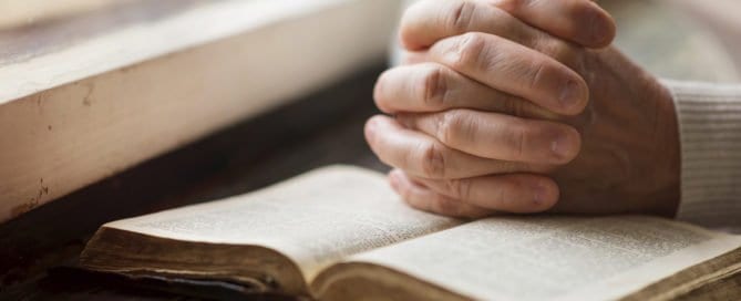 Person praying with hands clasped over an open Bible on a wooden table.