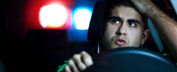Young man in police car with flashing red and blue lights in the background.