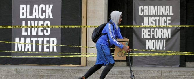 Person with backpack and scooter in front of protest banners about criminal justice reform.