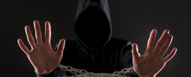 Person in black hoodie with hands raised, chained at the wrists, face hidden in darkness.